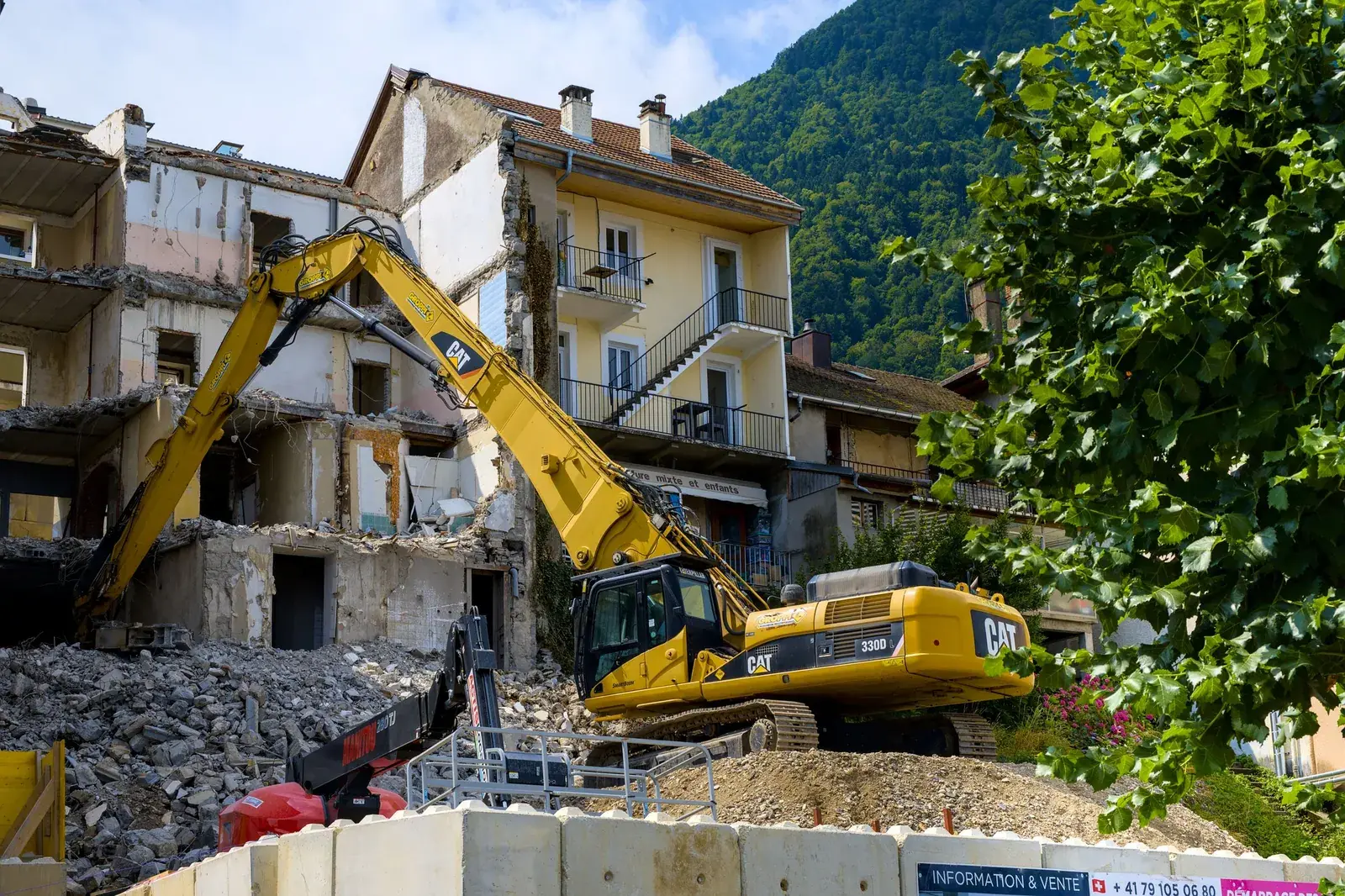 Excavator demolishing a building, with rubble in front and a green mountain behind