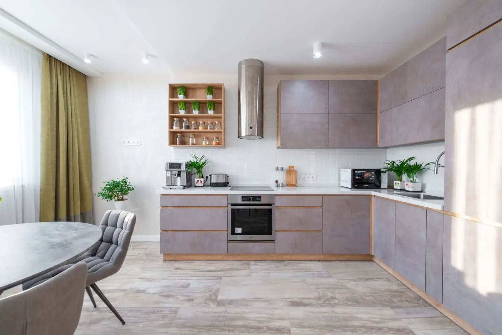 Kuzhinë moderne apartamenti me dollapë gri, aspirator inoks dhe tavolinë ngrënieje në dritën e ditës